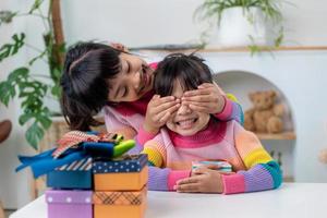 Little girl making present to little happy kid girl holding gift box,  closing eyes of cute smiling  excited with birthday surprise from her sister photo