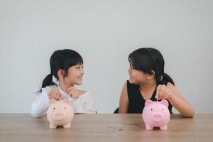 dos niñas asiáticas divirtiéndose para poner monedas en una alcancía juntas, niños ahorrando dinero para el concepto futuro foto