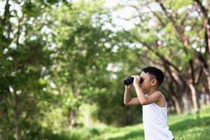 Little boy standing in a green grassy field scanning the surrounding woods with binoculars as he explores the countryside photo