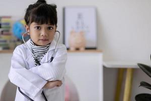 una niña asiática con anteojos y uniforme médico blanco sostiene un estetoscopio en la mano y quiere realizar un examen, juega al médico y al hospital. niño feliz actúa como médico que examina a alguien foto