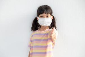 Coronavirus Covid-19 pm2.5.Online education.Little Asain girl wearing face mask show thumbs up for good and happy at home. Covid-19 coronavirus. Stay home. Social distancing.New normal behavior. photo