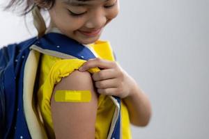 Asian young girl showing her arm with yellow bandage after got vaccinated or inoculation, child immunization, covid delta vaccine concept photo