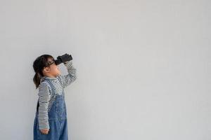 niña asiática mirando a través de binoculares en fondo blanco. concepto de exploración y aventura. foto