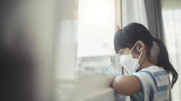 Little Asian girl wearing mask for protect pm2.5 and Covid-19. Sick child Little girls look at the window with longing on the street. Copy space. sadness child at home in isolation photo