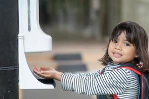 Child girls kid using automatic alcohol gel dispenser spraying on hands sanitizer machine antiseptic disinfectant, new normal life after Coronavirus COVID-19 pandemia. photo