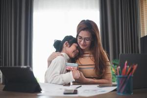 feliz día de la madre. una joven madre recibe felicitaciones de una emocionada y linda hija en casa, un niño le da a la madre una caja de regalo mientras trabaja en una laptop. concepto de vacaciones en familia foto