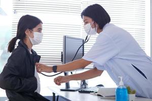 una doctora asiática que usa una máscara quirúrgica está revisando la frecuencia cardíaca del paciente infantil con un estetoscopio. foto