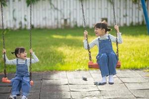 Two cute little sisters having fun on a swing together in a beautiful summer garden on a warm and sunny day outdoors. Active summer leisure for kids. photo