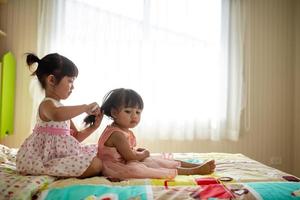 una niñita encantadora cepillando el cabello de su hermana mientras se sienta en la cama foto