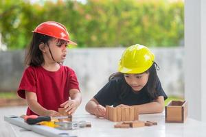 hermanos asiáticos niñas con sombreros de ingeniería construyendo casa con el juguete de madera. para aprender y mejorar el desarrollo, pequeño arquitecto. foto