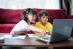 Two asian child girl students study online with teacher by video call together. Siblings are homeschooling with computer laptop during quarantine due to Covid 19 pandemic. photo