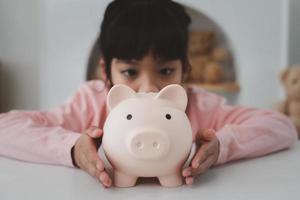 niña asiática ahorrando dinero en una alcancía, aprendiendo a ahorrar, los niños ahorran dinero para la educación futura. concepto de dinero, finanzas, seguros y personas foto