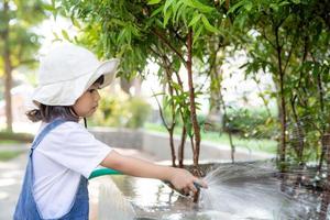 niña asiática vertiendo agua en los árboles. niño ayuda a cuidar las plantas con una regadera en el jardín. foto