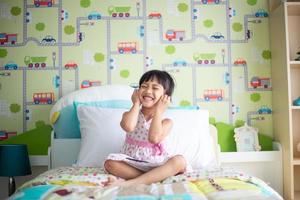 niños asiáticos usando auriculares para escuchar música por teléfono inteligente en la cama en su dormitorio decorado foto