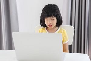 una pequeña estudiante asiática estudia en línea usando una computadora portátil en casa foto