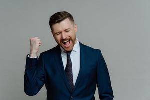Positive businessman clenches fist exclaims from joy hooray celebrates success dressed formally satisfied after successful business deal isolated over grey background. Congratulations I did it photo