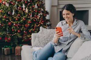 Happy female in earbuds holding smartphone, talking with family or friends online at Christmas time