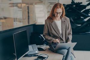 Female office worker sitting on top of desk while working with project files in modern laptop photo