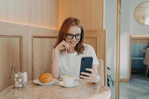 Young positive redhead woman in earphones sitting in cafe and talks online on smartphone photo