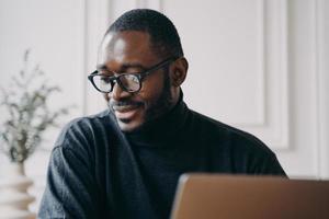 un hombre afroamericano pensativo se sienta en el escritorio de su casa, ve una clase de video en línea o un seminario web en la computadora foto