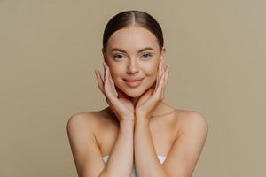 Photo of satisfied refreshed young woman touches face gently has clean healthy skin wears minimal makeup wrapped in bath towel shows bare shoulders isolated over brown background. Beauty concept