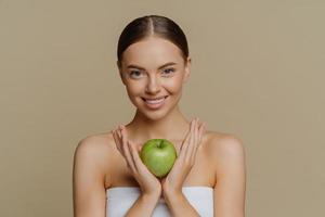 Beautiful young female model with perfect fresh smooth skin holds green apple containing much vitamins stands indoor wrapped in bath towel isolated over brown background. Natural care cosmetics photo