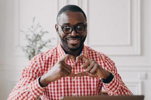 Happy african man gesturing heart symbol shape with hands during video call with family photo