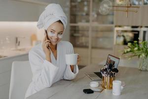 Happy young European woman undergoes beauty procedures after morning shower wears hydrogel moisturising patches dressed in white bathrobe drinks aromatic coffee talks via mobile phone poses indoor photo