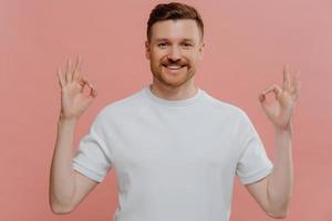 Young satisfied bearded man makes okay gesture with both hands expresses agreement approves something praises nice work says well done dressed in casual white t shirt isolated on pink studio wall photo