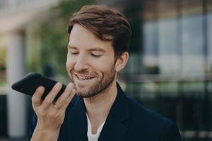 Handsome smiling neat bearded office worker talking on speakerphone standing outside photo