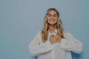 Happy thankful woman with blond wavy hair in white oversize shirt gesturing appreciation and love photo