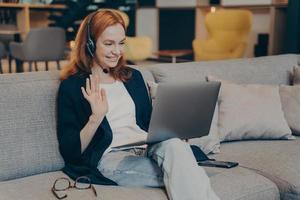 Young woman using laptop and headset for internet video call waving hand in greeting gesture photo