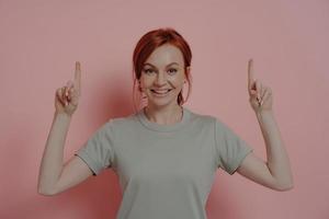 Happy young caucasian woman pointing upwards with index fingers and smiling cheerfully photo