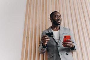 emocionado hombre de negocios afroamericano usando teléfono inteligente y tarjeta de crédito para comprar en línea foto