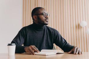 Thoughtful african businessman sits at desk with pen and agenda looks away with pensive expression photo