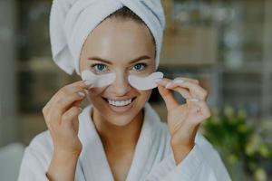 Headshot of happy young blue eyed woman with minimal makeup takes off beauty patches enjoys good effect has healthy well cared skin white perfect teeth wears bathrobe and wrapped towel on head photo