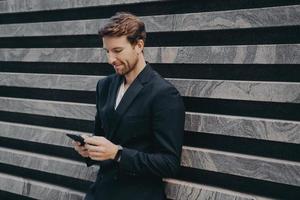 Pensive young office worker in formal stylish suit standing outdoor holding mobile phone in hands photo