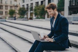 Young satisfied businessman in stylish suit sitting on stairs outside with laptop and coffee to go photo