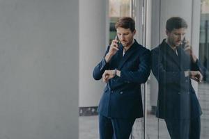 Pensive businessman in suit talking on mobile phone standing outside, looking at his hand watch photo