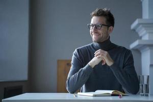 Pensively smiling male entrepreneur sitting at table in front of open agenda with folded hands photo