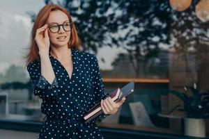 Female entrepreneur carries tablet smartphone and organizer poses near window of restaurant photo