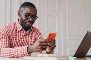 Young handsome african male executive in glasses sitting at office table and using mobile phone photo