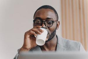 Attractive confident Aframerican man company CEO drinking coffee while looking at laptop screen photo