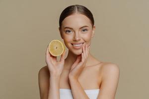 Healthy glad young woman poses with bare shoulders indoor wrapped in bath towel holds slice of lemon smiles tenderly has minimal natural makeup isolated over brown background. Skin care concept photo