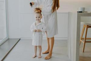 Faceless mother makes pony tail from daughters curly hair uses comb. Cute little three year old girl in white bathrobe after taking shower poses near mom at home. Children parenthood beauty time photo