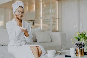 Horizontal shot of pretty Caucasian woman applies facial cream looks thoughtfully aside holds bar of cosmetic product dressed in bathrobe and towel sits on comfortable sofa over home interior photo