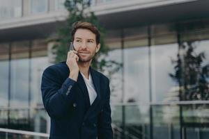 Male entrepreneur talks via phone solves urgent problems poses near business center photo