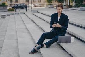 Young confident man in stylish suit using smartphone while sitting on steps outside with laptop photo