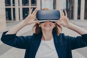 joven empresaria pelirroja sonriente probando gafas vr portátiles en las calles de la ciudad foto