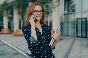 Cheerful redhead European woman talks on modern mobile phone has glad expression